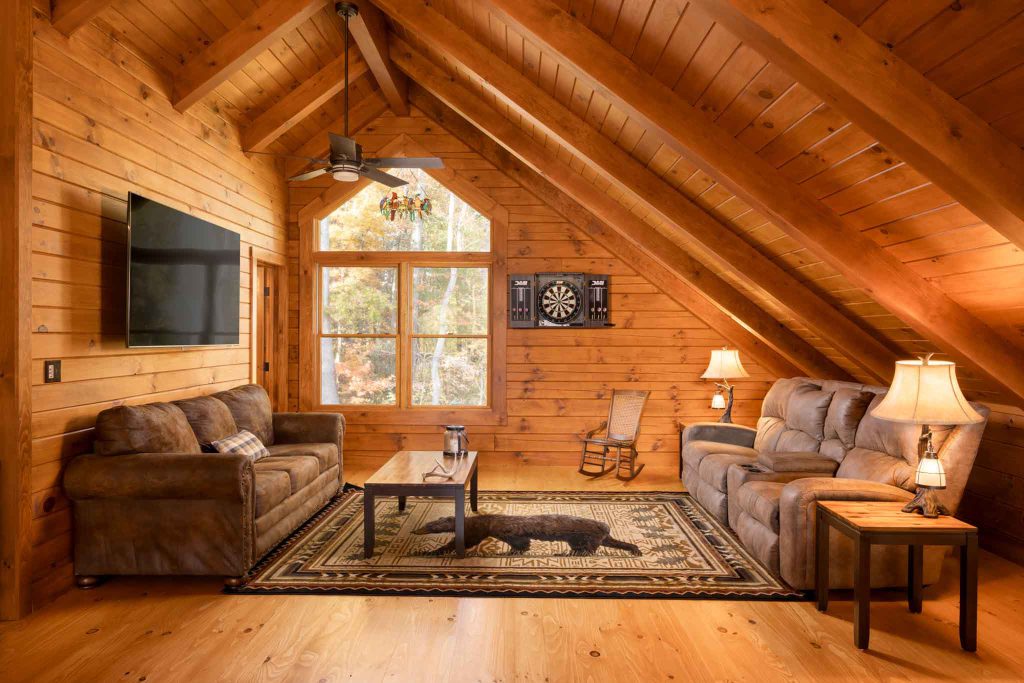 A cozy wooden living room featuring a high ceiling with exposed beams, a comfortable brown sofa, a rocking chair, and a coffee table on a patterned rug. A large window lets in natural light and offers a view of trees outside.