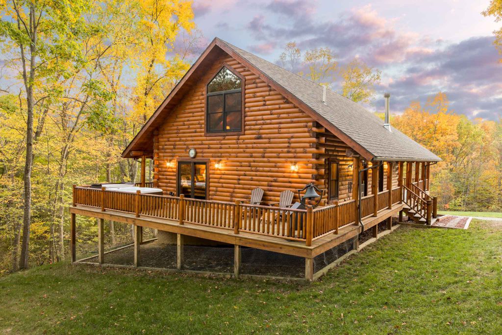 A beautifully constructed log cabin nestled in a forest with vibrant autumn foliage. The cabin features a spacious wooden deck with chairs, large windows, and a mountain view against a dramatic sky. Ideal for a cozy retreat.