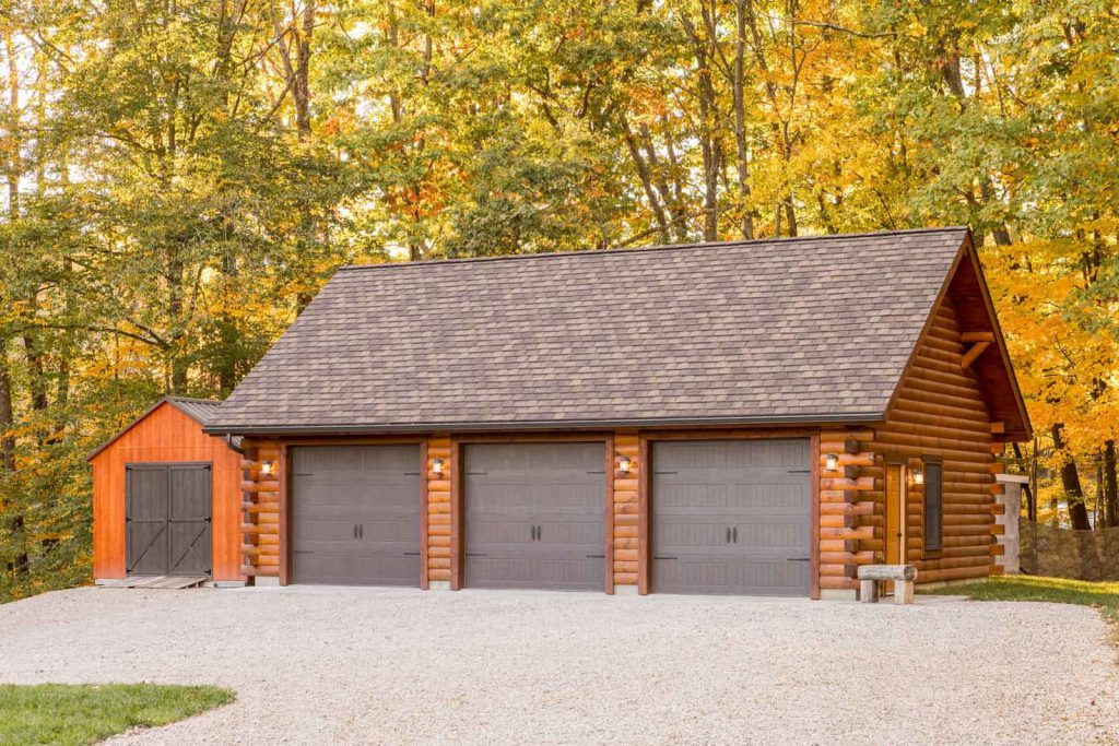 A spacious log cabin garage with three large doors sits beside a smaller shed, surrounded by trees displaying vibrant autumn foliage. A gravel driveway leads up to the garage, enhancing the rustic charm of the setting.