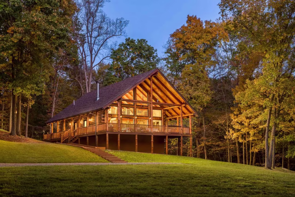 A charming wooden lodge with a steeply pitched roof is nestled among tall trees, illuminated by warm lights at dusk. The porch showcases sturdy railings, inviting a serene atmosphere in a woodland setting.