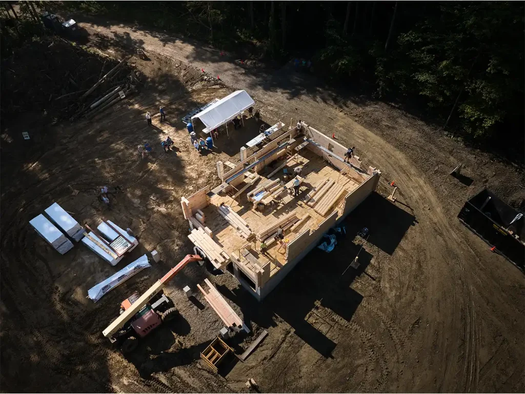 Aerial view of a log home package being assembled at a wooded construction site, with stacked timbers, wall sections, equipment, and workers.
