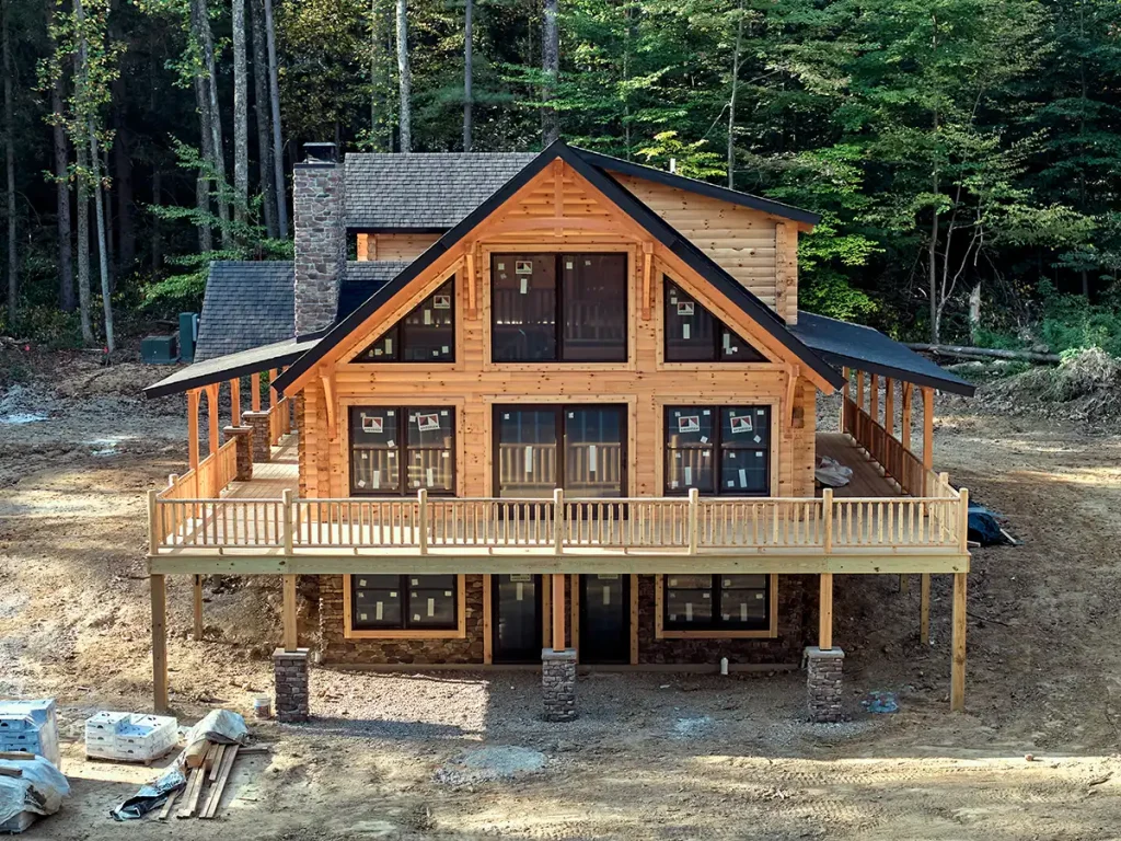 New log home under construction in a wooded setting, featuring expansive gable windows, a stone chimney, wraparound deck, and walkout basement.