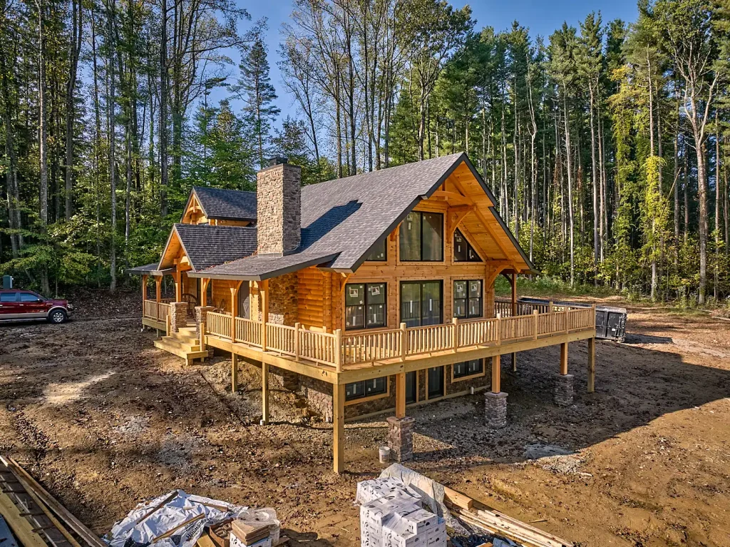 Hochstetler spec log home under construction among towering pine trees, featuring a stone chimney, expansive gable windows, and wraparound deck.