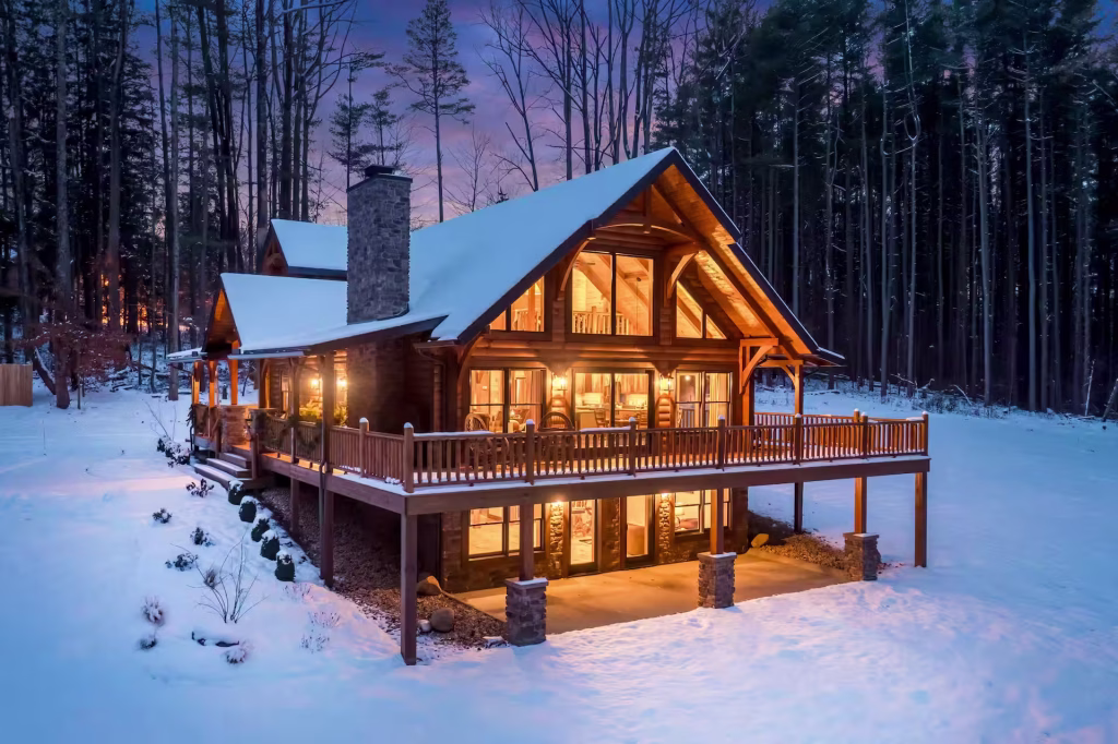 Angled view of the Hochstetler Log Home showing wraparound deck, stone chimney, and covered walkout patio in winter