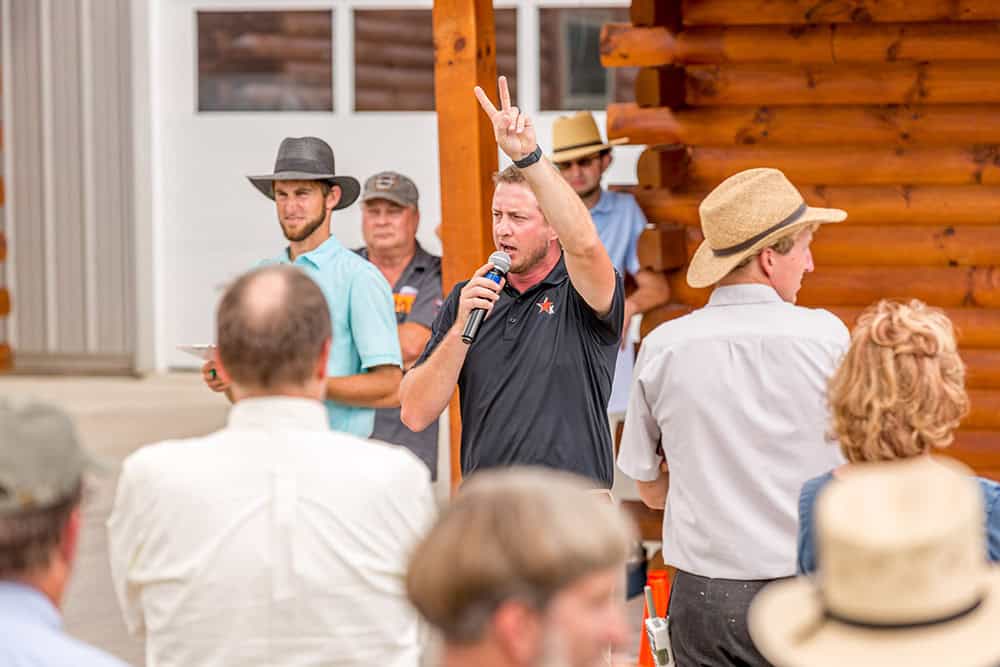 Man speaking at vendor registration event with audience and log home background.