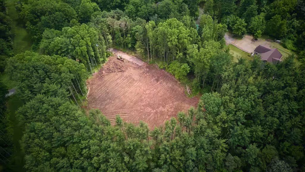Aerial view of a cleared building pad cut into heavy woods, bare earth in the centre and a neighbouring house at the edge of the trees