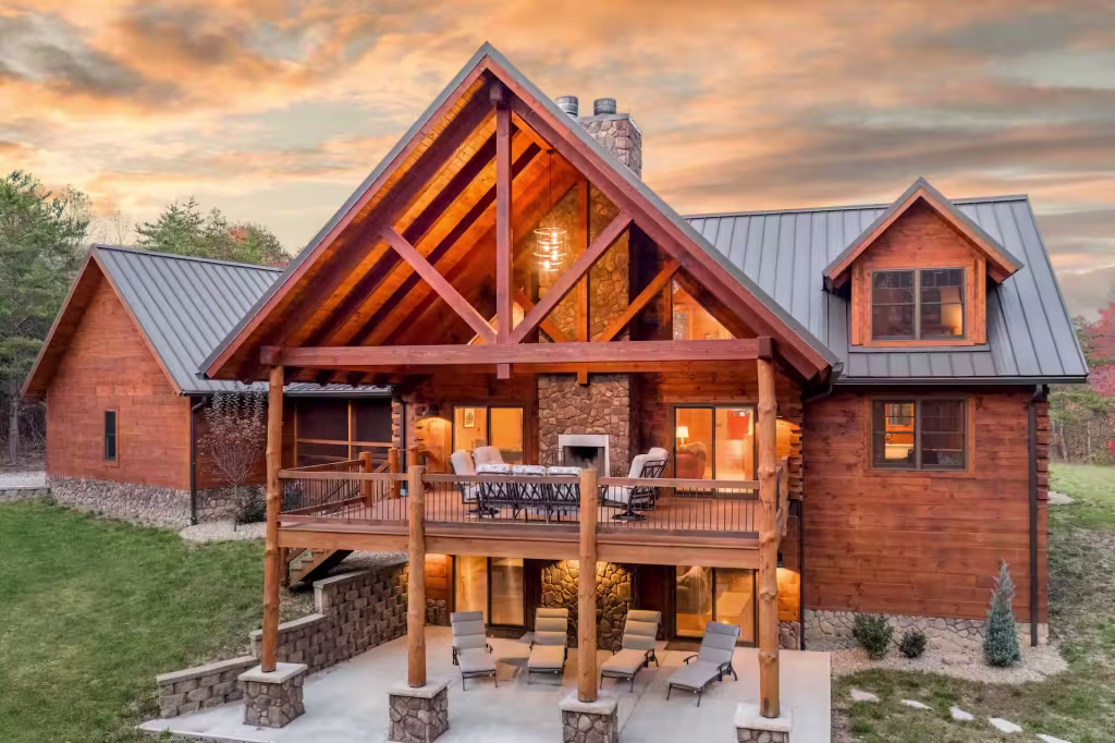 Rear exterior at dusk showing a two-story wall of windows under a timber-truss gable, an upper deck, and a lower patio with loungers.