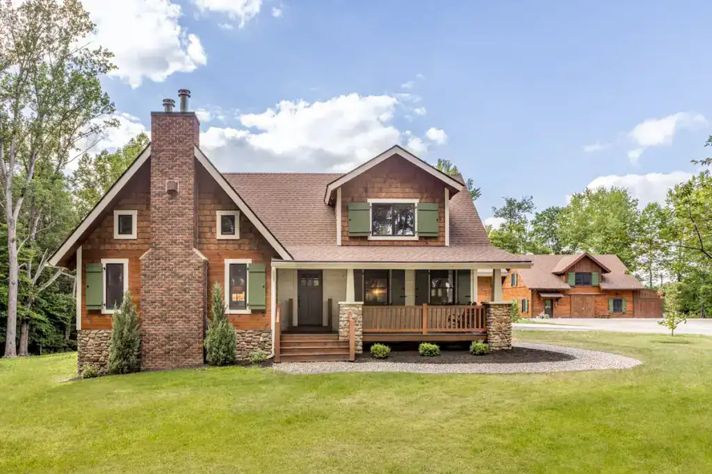 Historic-inspired Ohio log home with green shutters, brick chimney, covered porch and matching barn.
