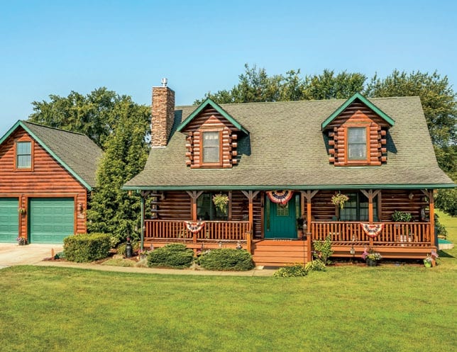 Beautiful log cabin with green roof and front porch, surrounded by lush lawn and trees.