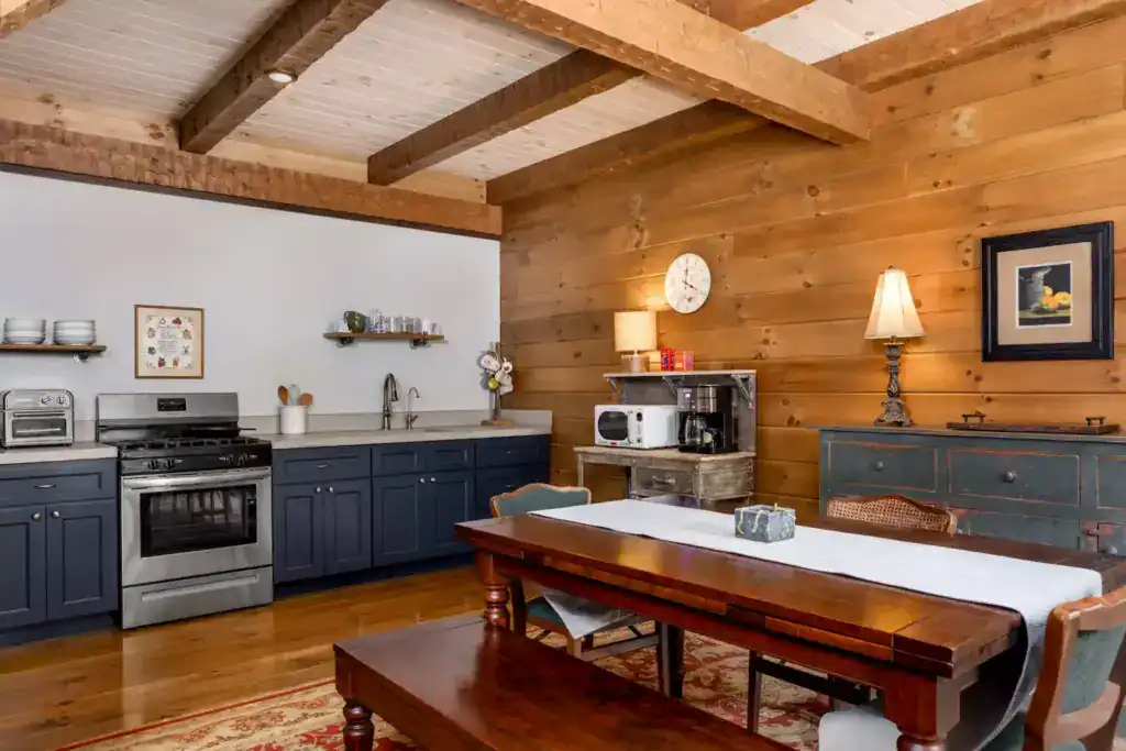 Kitchen and dining area with slate blue cabinets, a stainless range, pine plank walls, exposed beams and a long wood dining table