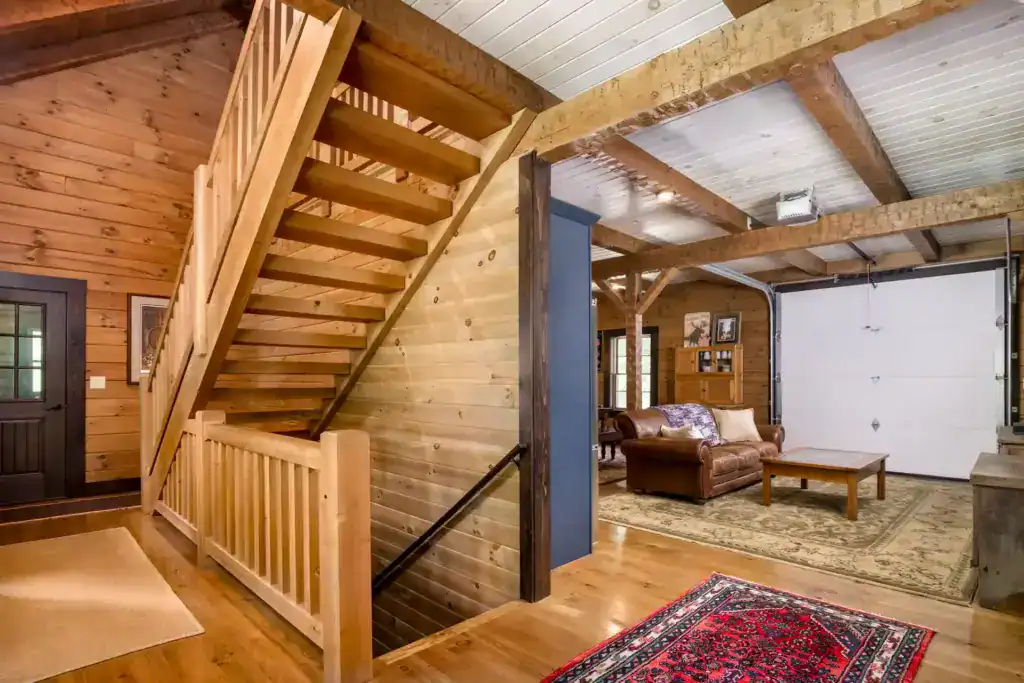 Open wood staircase with a pine railing beside a living area with a leather sofa and a converted garage door, under exposed beams