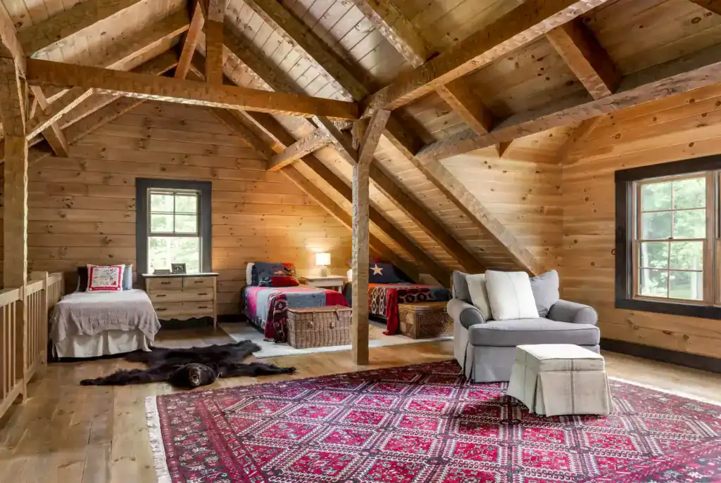 Sleeping loft under exposed timber trusses with three beds, pine plank walls, a red rug and a gray reading chair