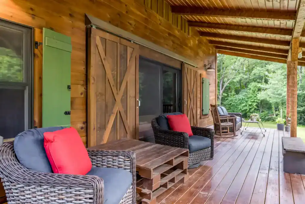 Covered back porch with sliding barn doors, green shutters, wicker chairs with red pillows and a pallet coffee table