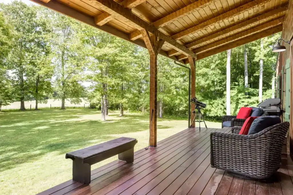 Wide covered porch with timber posts and a plank ceiling overlooking a lawn and trees, with wicker chairs, a bench and a telescope