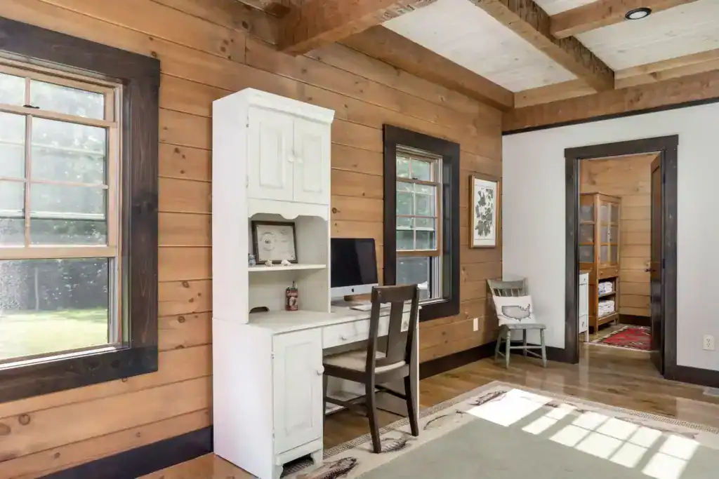 Bedroom corner with pine plank walls, dark-trimmed windows, a white desk hutch and exposed ceiling beams