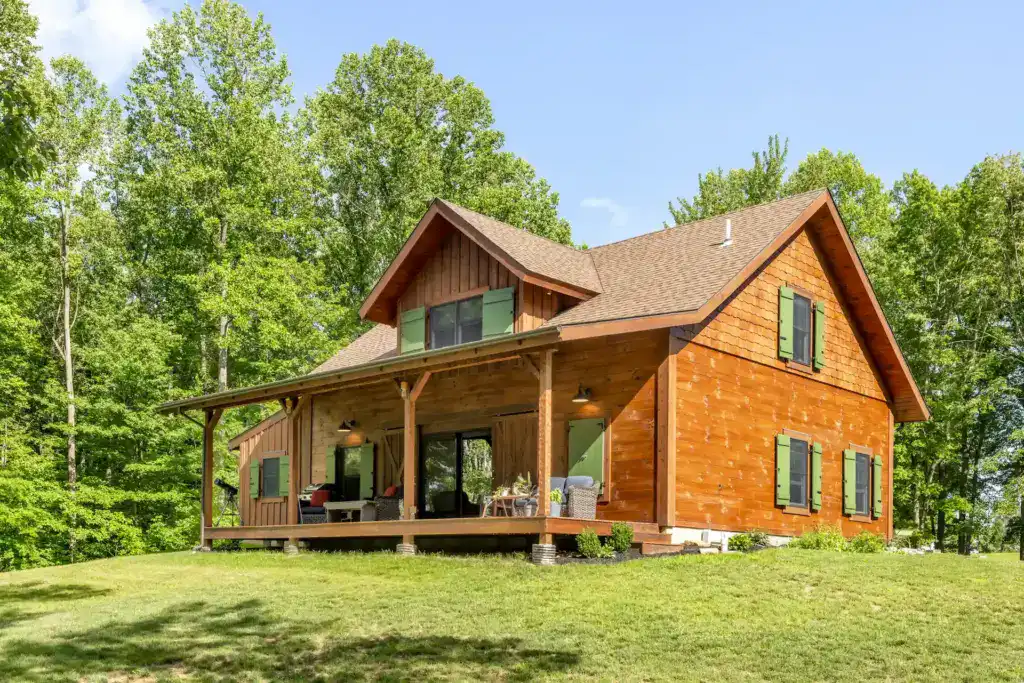 Side view of a wood-sided barn with a wraparound covered porch, green shutters, shingled gable and a dormer on a green lawn