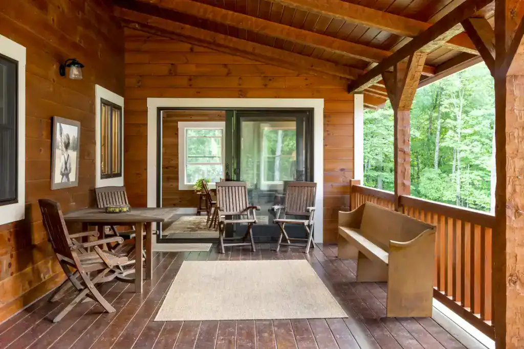 Covered back porch with a plank ceiling, timber posts, wooden chairs and table, a church-pew bench and a view of the woods