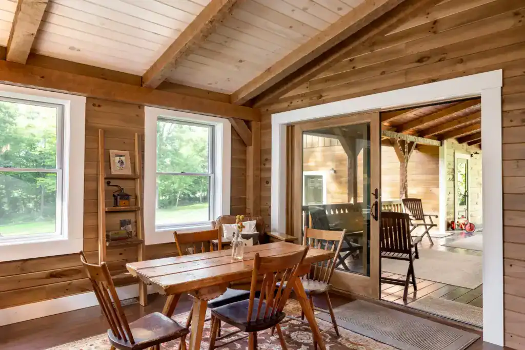 Dining area with exposed timber beams, wood plank walls, a farmhouse table and chairs, and glass doors to the covered porch