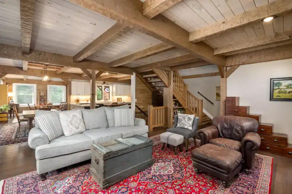 Living room with a timber-frame ceiling, open staircase, a leather armchair and a vintage trunk coffee table on a red rug