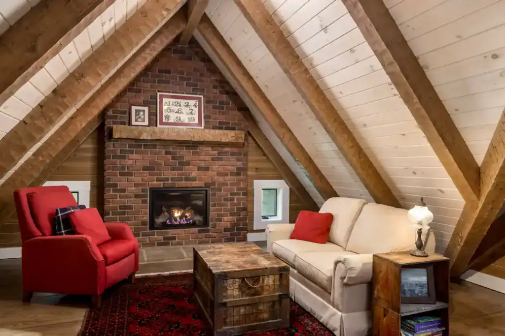 Upstairs loft under exposed timber rafters with a brick gas fireplace, a red armchair and a cream sofa