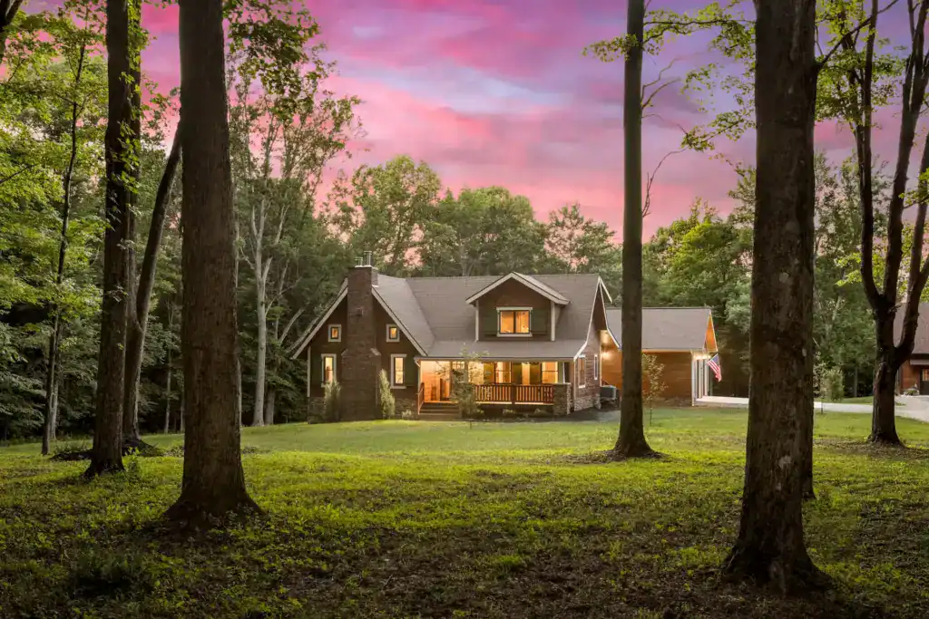Log home glowing at dusk under a pink sky, framed by tall trees, with lights on in the front porch and windows